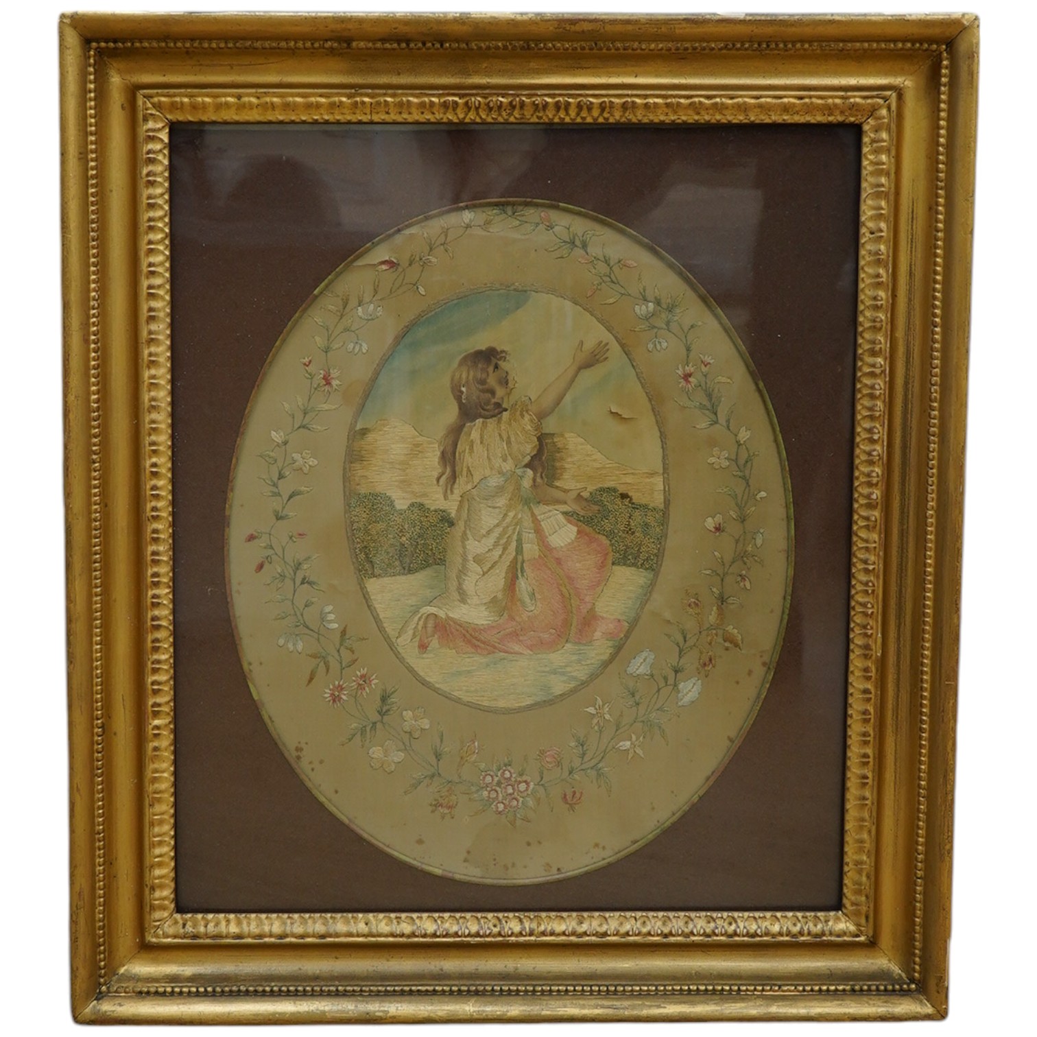 A Regency oval silkwork embroidered panel of a girl in a landscape with a floral embroidered border and gilt frame, 35cm high x 28cm wide. Condition - the silk background has torn in places